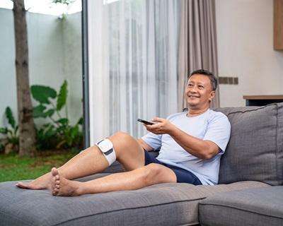 A man sitting on a couch, using OMRON Knee TENS HV-F710-M while watching TV.